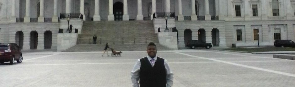 Person in a black vest and tie stands in front of a large neoclassical government building with steps.