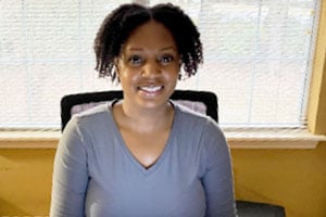 Smiling woman seated at a desk in a bright office with a window behind her.