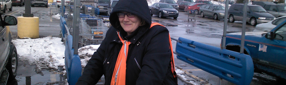 Smiling woman in a black hooded coat with an orange scarf beside a blue barrier in a snowy parking lot.