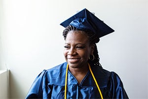Smiling graduate woman in blue cap and gown with yellow cords.
