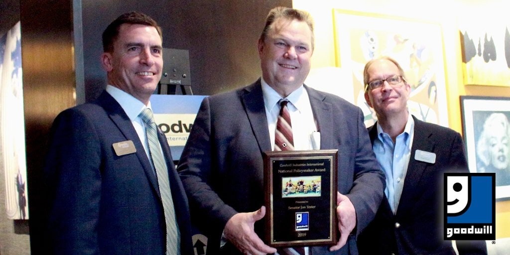 Three men in business suits pose with a national policymaker award plaque at a Goodwill event, with a Goodwill logo in the corner.