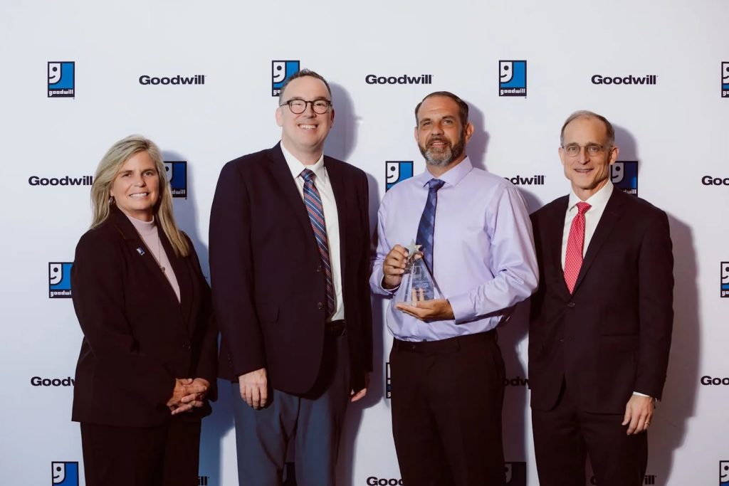 Four professionals in business attire pose with an award at a Goodwill event.