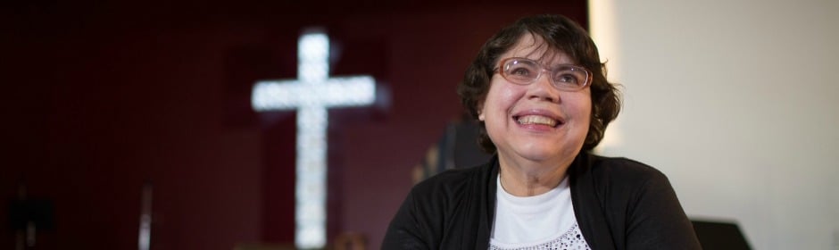 Smiling woman with short dark hair and glasses sits indoors, with a lit cross in the background.