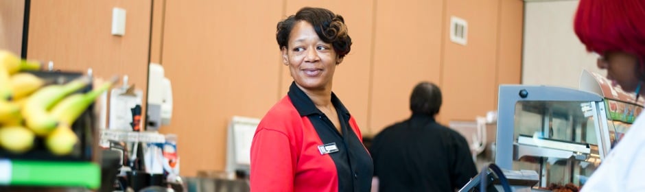 Smiling store associate in a red uniform at the checkout, with a fruit display in the foreground.