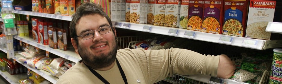 Smiling man with glasses reaching into a shopping cart in a grocery store aisle lined with cereal boxes.