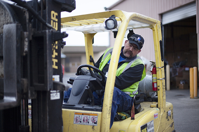 Forklift operator wearing a high-visibility vest and cap in a yellow forklift cab at an industrial yard.
