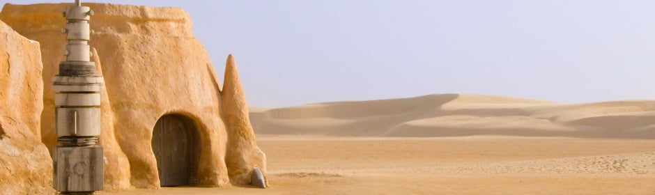 Desert scene with a mud-brick hut, arched doorway, and a weathered metal device beside rolling sand dunes.