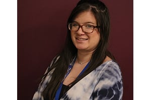 Smiling woman with long dark hair and glasses wearing a blue and white tie-dye top against a maroon background.