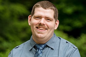 Smiling man in blue shirt outdoors with greenery in the background