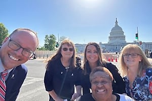 Group of five smiling adults taking a selfie in front of the U.S. Capitol on a sunny day.
