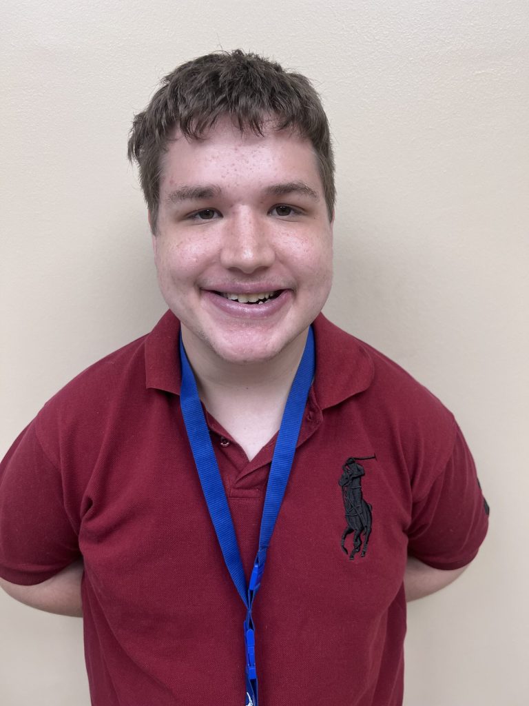Young man with short dark hair, smiling, wearing a burgundy polo and blue lanyard against a beige wall.