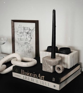 Black taper candle in a marble holder atop design books, with a framed sketch and decorative objects on a dark table.