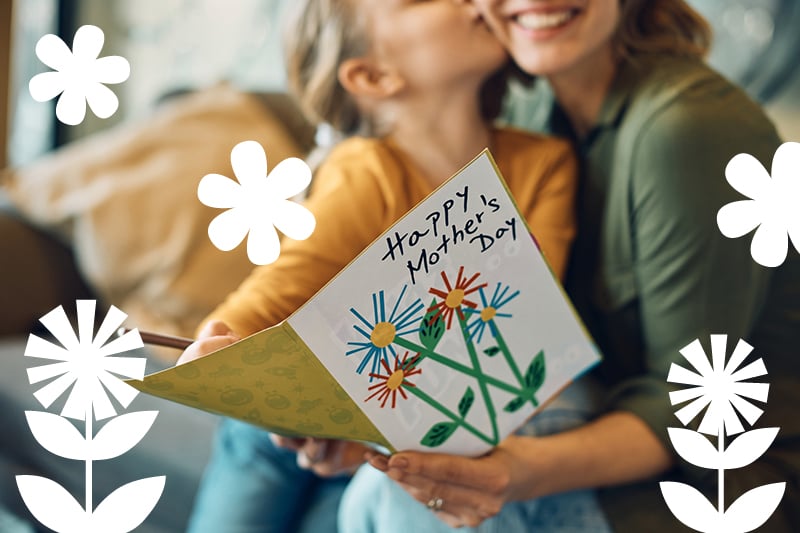 Mother and child sharing a handmade 'Happy Mother's Day' card with colorful flowers, framed by white decorative petals.
