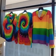 Rainbow tie-dye and striped garments on hangers in a storefront window.