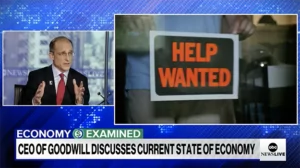 News broadcast screenshot with a man speaking and a prominent 'HELP WANTED' sign in the foreground.