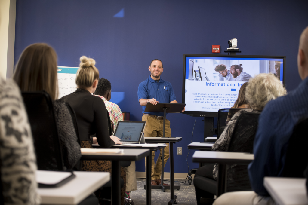 Trainer in a blue polo presenting to adult learners in a classroom with a large informational slide on a screen.