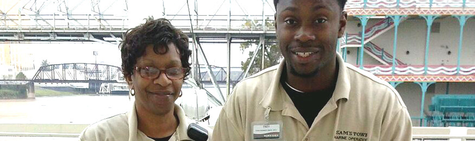Two uniformed staff members stand outdoors at a riverfront exhibit, smiling at the camera.