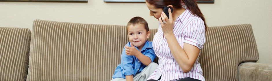 Mother sits on a beige striped couch, talking on a smartphone while her smiling son gives a thumbs up.