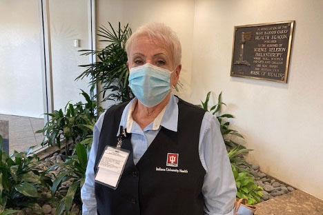 Elderly healthcare worker wearing a blue surgical mask and navy vest with an ID badge in a plant-filled hospital lobby.