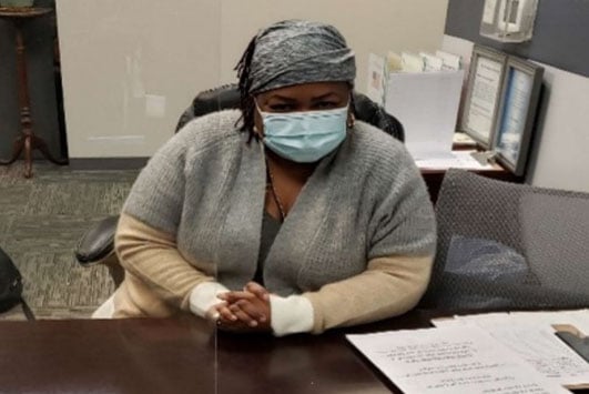 Woman wearing face mask and headscarf sits at a desk with documents in an office.