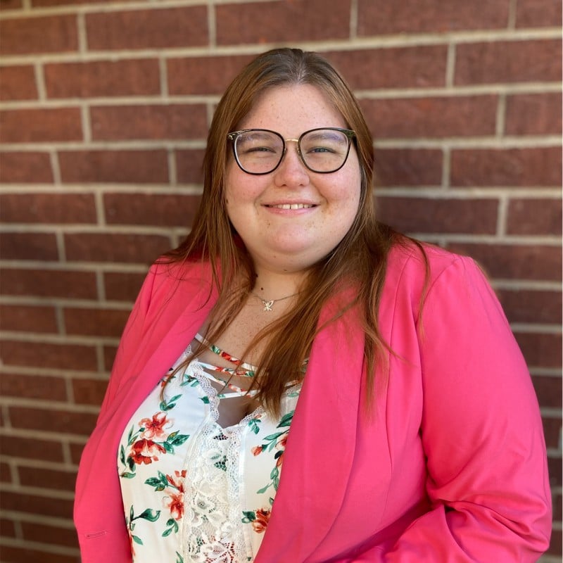 Smiling woman with long brown hair and glasses, wearing a pink blazer over a floral blouse, posed against a brick wall.
