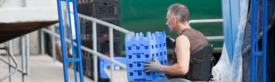 Man in a wheelchair carrying blue plastic crates in a loading dock.