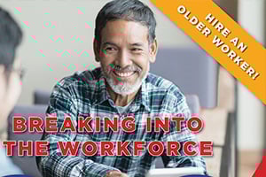 Older man with gray hair and a beard in a plaid shirt smiles during a discussion in a casual office setting.