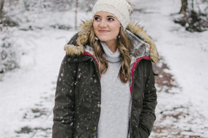 Woman in a dark winter parka with a fur-lined hood and knit beanie standing on a snowy path, smiling.