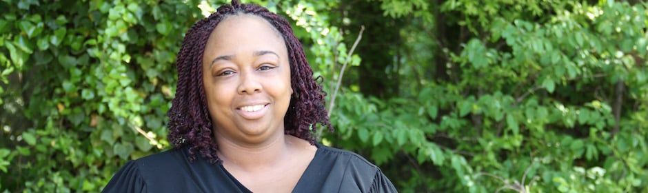 Smiling woman outdoors with lush green shrubs in the background.