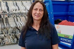 Woman with long dark hair in a navy polo standing near blue recycling bins in a storage area.