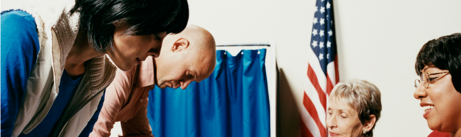 Diverse group of adults gathered near a voting booth, with an American flag in the background.