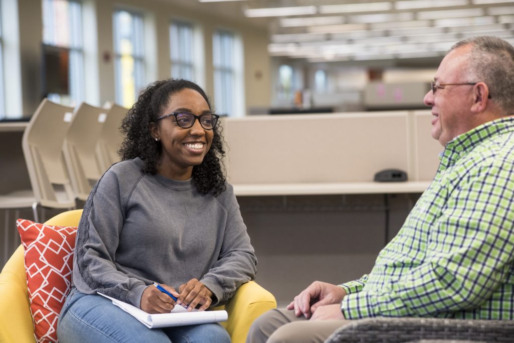 Smiling woman with glasses and notebook conversing with a man in a green plaid shirt in a bright, open office.