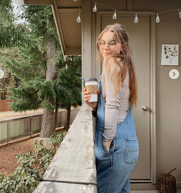 Young woman with glasses wearing denim overalls stands on a balcony railing, holding a takeaway coffee outdoors.