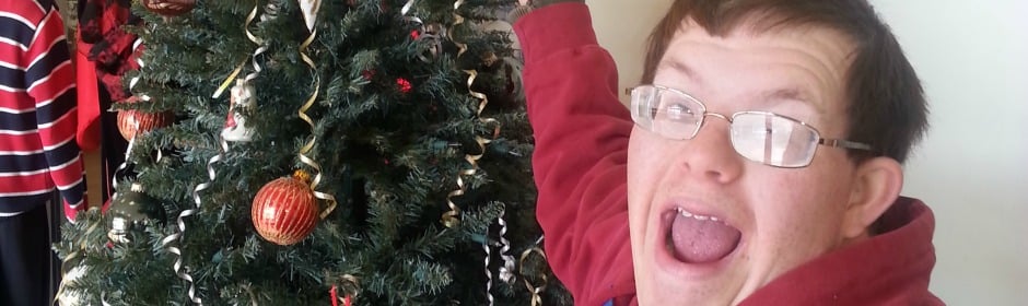 Person in a red hoodie posing excitedly beside a Christmas tree decorated with ornaments.