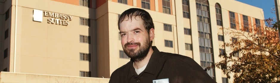 Bearded hotel staff member in a dark polo with a name badge stands outside an Embassy Suites hotel during daytime.