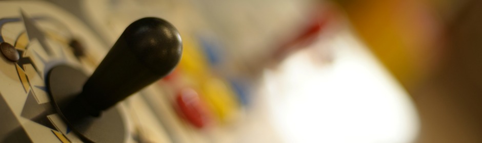 Close-up of a black joystick on a control panel with blurred colored dials in the background.
