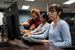 Three people at computer workstations in a lab, with an instructor guiding two students.
