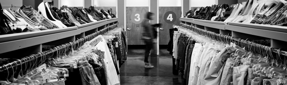 Blurred shopper walks between long racks of clothes in a grayscale thrift store, back wall shows numbered doors 3 and 4.