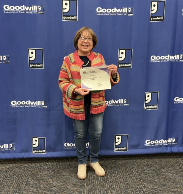 Smiling woman in a colorful striped cardigan holds a certificate in front of a Goodwill backdrop.