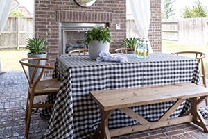 Outdoor dining setup on a brick patio with a brick fireplace, featuring a black-and-white checkered tablecloth and potted greenery.