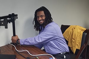 Smiling man with dreadlocks in a purple shirt sits at a desk with a laptop.