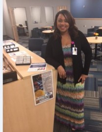 Smiling woman in a colorful striped dress and black cardigan stands at a wooden reception counter in a busy lobby.