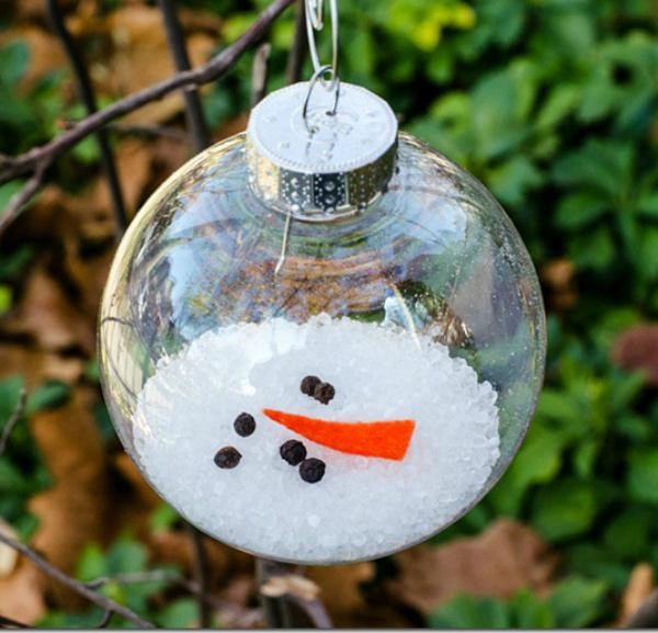 Glass ornament ball filled with snow forming a tiny snowman with coal eyes and a carrot nose, hanging outdoors.