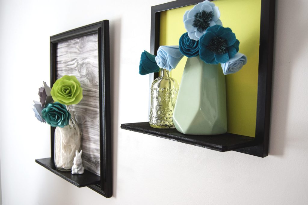 Two wall-mounted black-framed shelves display vases and handmade fabric flowers in teal, blue, and lime.