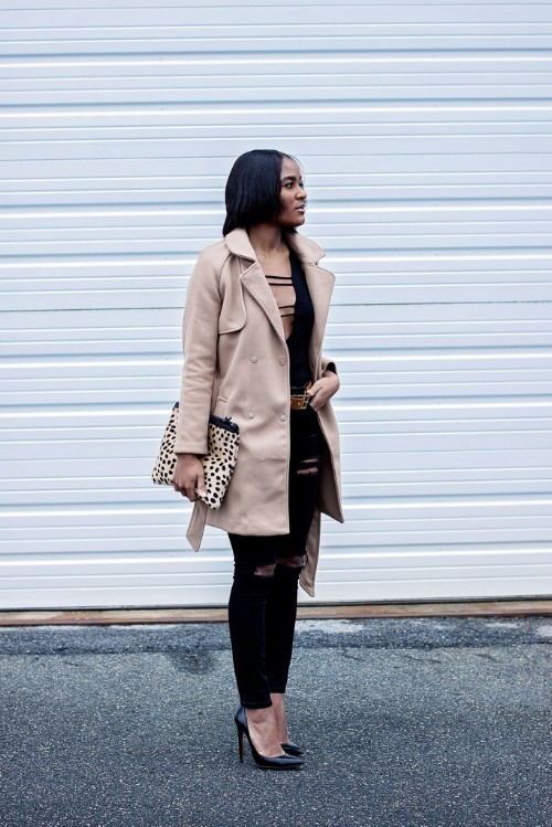 Stylish woman in a beige trench coat, black ripped jeans, and a leopard-print clutch against a light metal backdrop.