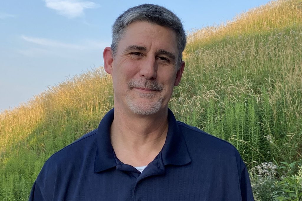 Portrait of a middle-aged man with short gray hair and stubble, wearing a navy polo, standing in a sunlit grassy field.
