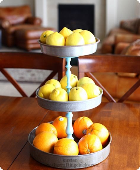 Three-tier metal fruit stand filled with lemons on the top two levels and oranges on the bottom, set on a wooden table.