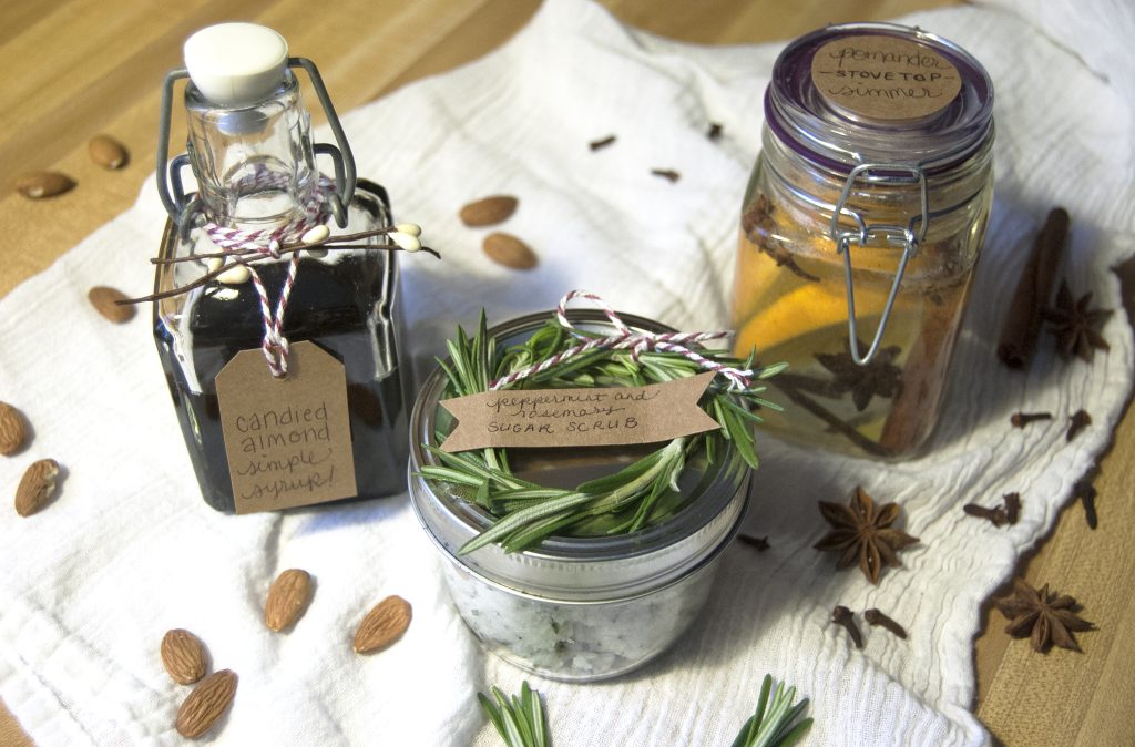 Assorted handmade gifts on a white cloth: rosemary peppermint sugar scrub, candied almond syrup, and a citrus-infused jar.