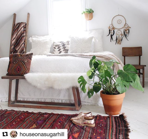 Boho bedroom with white bedding, a wooden bench, patterned pillows, fluffy throw, red woven rug, dreamcatcher wall decor, and a potted monstera.