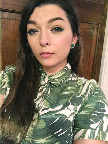 Woman with long dark hair wearing a green tropical leaf-pattern shirt and turquoise earrings, indoors.
